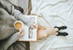 woman holding a cup of coffee at right hand and reading book on her lap while holding it open with her left hand in a well-lit room