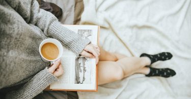woman holding a cup of coffee at right hand and reading book on her lap while holding it open with her left hand in a well-lit room
