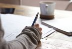 person writing on brown wooden table near white ceramic mug