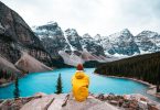 person in yellow hoodie sitting on rock near lake and snow covered mountain during daytime