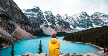 person in yellow hoodie sitting on rock near lake and snow covered mountain during daytime